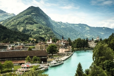 Vue de la ville avec rivière, bateaux et montagnes environnantes en arrière-plan.