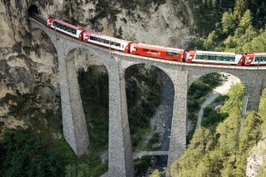 Red train crossing a high stone bridge in a mountain landscape.