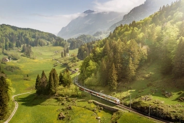 Eine Bahn fährt durch eine grüne, bergige Landschaft mit Wäldern und Wiesen.