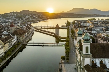 Sunset over a city with a bridge and river, mountains in the background.