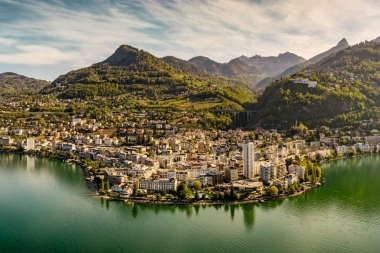 Città sul lago con colline boscose e montagne sullo sfondo.