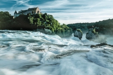 Cascata con castello sullo sfondo, circondata da vegetazione lussureggiante.