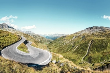 Windy mountain road in a green, hilly landscape under a clear, blue sky.