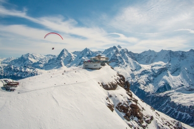 A snowy mountain landscape with a building on a peak and a paraglider in the sky.