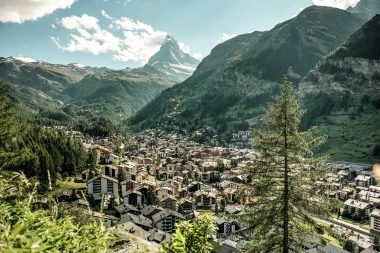 View of an alpine valley with a town, surrounded by green mountains and a prominent mountain peak in the background.