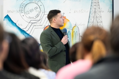 A person speaking into a microphone in front of a colorful mural featuring the Eiffel Tower and a postage stamp.