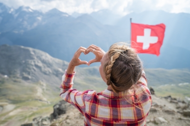 A woman forms a heart with her hands in front of a Swiss flag and mountain backdrop.