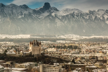 Eine winterliche Stadt bei Nacht, beleuchtete Gebäude, schneebedeckte Dächer und Berge im Hintergrund.