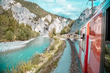 Roter Zug fährt entlang eines blauen Flusses in einem Bergtal.