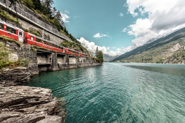 Red train travelling along the edge of a mountain by a lake.