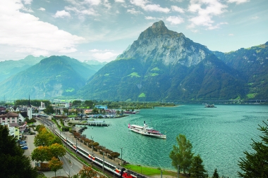 Mountain landscape with lake, train, and ship in the foreground.