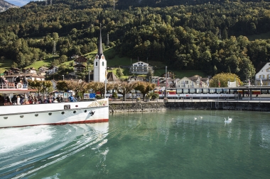 A ferry on a lake, with a church and houses in a green mountain landscape in the background.