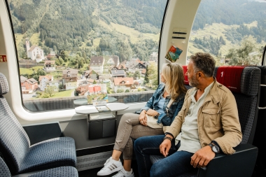 A couple sits on a train and enjoys the view of a mountain village from the panoramic window.