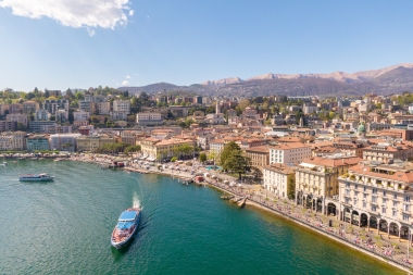 Vista aerea del Lago di Lugano con battello turistico e montagne sullo sfondo in una giornata di sole.