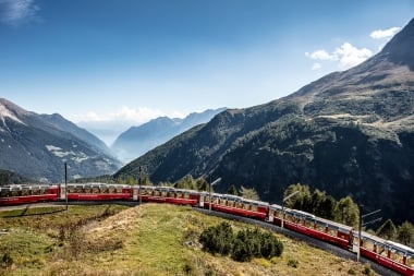Der Bernina Express fährt entlang einer grünen Wiese, im Hintergrund ist ein Bergpanorama zu sehen.