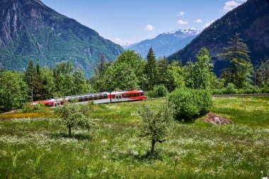 Der Mont-Blanc Express fährt durch eine grüne Landschaft, im Hintergrund ist ein Alpenpanorama zu sehen.