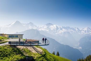 People are playing the alphorn on a viewing platform with a mountain panorama in the background.
