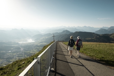 Two hikers are walking down the mountain path, enjoying views of a lake and the mountains.