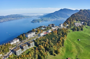 An aerial view shows a hotel complex situated on a mountain, surrounded by woodland, with a lake in the background.