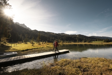 Zwei Personen laufen auf einem Steg an einem See, der von einem herbstlichen Wald umgeben ist.
