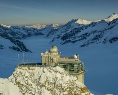 Eine Sternwarte auf einem schneebedeckten Berggipfel mit Berglandschaft im Hintergrund.