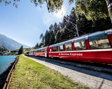 Un train rouge Bernina Express circule le long d'un lac bleu dans un paysage montagneux sous le soleil.