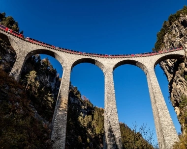 Un train rouge circule sur un viaduc en pierre dans un paysage rocheux sous un ciel bleu.