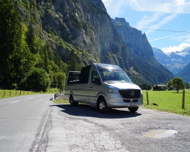 Ein silberner Transporter steht auf einer Landstrasse vor einer beeindruckenden Bergkulisse.