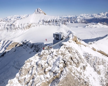 A snowy mountain landscape with a modern building and a group of people on the summit.