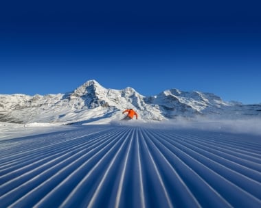 Skifahrer in orange auf frisch präparierter Piste vor schneebedeckten Bergen.