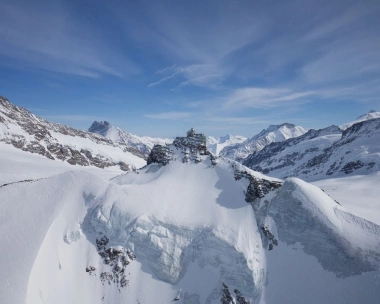 A snowy mountain landscape with a building on a peak under a blue sky.