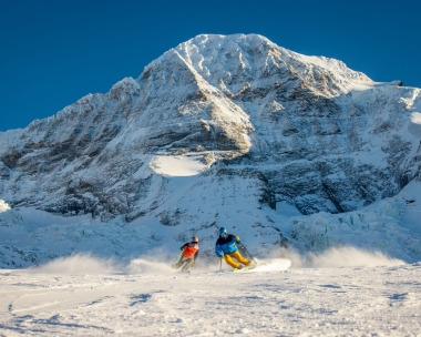 Zwei Skifahrer auf einer verschneiten Piste vor einem grossen, schneebedeckten Berg.