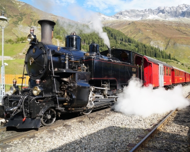 Eine schwarze Dampflokomotive zieht rote Personenzüge durch eine bergige Landschaft.