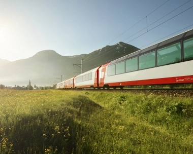 Un treno panoramico rosso attraversa un pittoresco prato verde con fiori colorati, sullo sfondo si ergono montagne maestose sotto un cielo azzurro splendente.