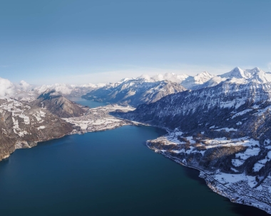 Luftaufnahme eines verschneiten Gebirges mit einem See im Vordergrund.