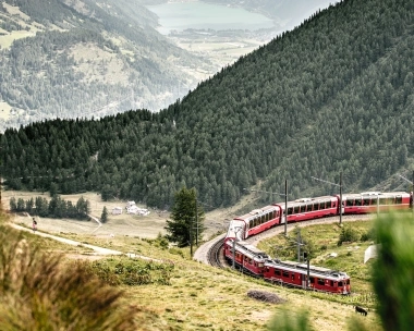A red train winds through a mountainous landscape with forests and a view of a lake in the distance.