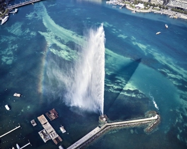 Aerial view of a large fountain in the water, surrounded by boats and a shore with buildings.