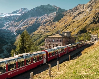 Un treno panoramico rosso attraversa un paesaggio alpino con alte montagne e una casa di pietra.