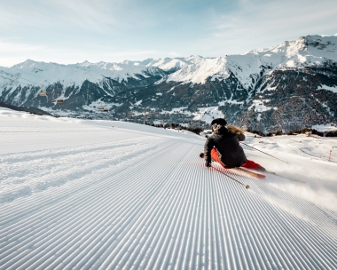 Eine Skifahrerin auf einer präparierten Piste mit schneebedeckten Bergen im Hintergrund.