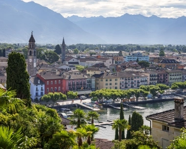 Eine Stadtansicht mit Kirchtürmen, bunten Gebäuden und einem See im Vordergrund, umgeben von bewaldeten Hügeln und Bergen.