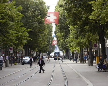 Une rue animée avec des arbres, des drapeaux suisses et un tramway qui passe.