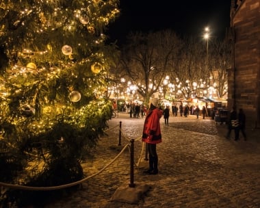 Eine Person steht vor einem grossen, beleuchteten Weihnachtsbaum auf einem festlichen Platz bei Nacht.