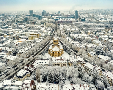 Aerial view of a snowy city with a church in the center and modern buildings in the background.