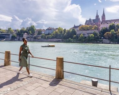 A woman walks along the riverbank, with historic buildings and a bridge in the background.