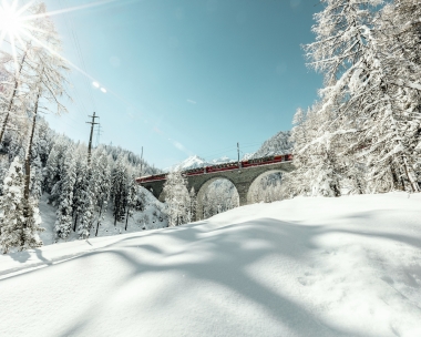 Eine rote Bahn überquert eine steinerne Brücke in einer verschneiten Berglandschaft.