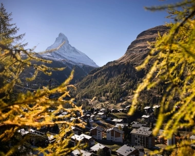A mountain landscape with a cluster of houses in the valley, a snow-capped peak in the background, framed by autumn-colored branches.