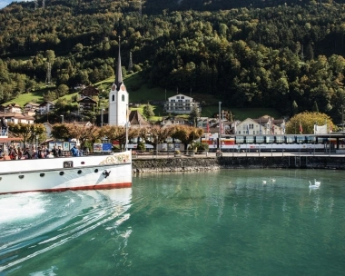 A ferry on a lake, with a church and houses in a green mountain landscape in the background.