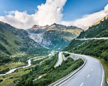 Une route de montagne sinueuse traversant une vallée verte vers des sommets montagneux imposants, avec un ciel nuageux en arrière-plan.