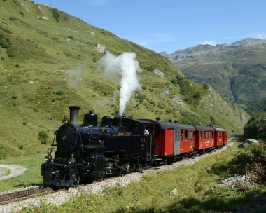 Eine schwarze Dampflokomotive zieht rote Personenzüge durch eine grüne Berglandschaft.