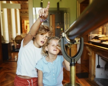 Deux enfants regardent un objet à travers un grand télescope dans une pièce avec un plancher en bois.